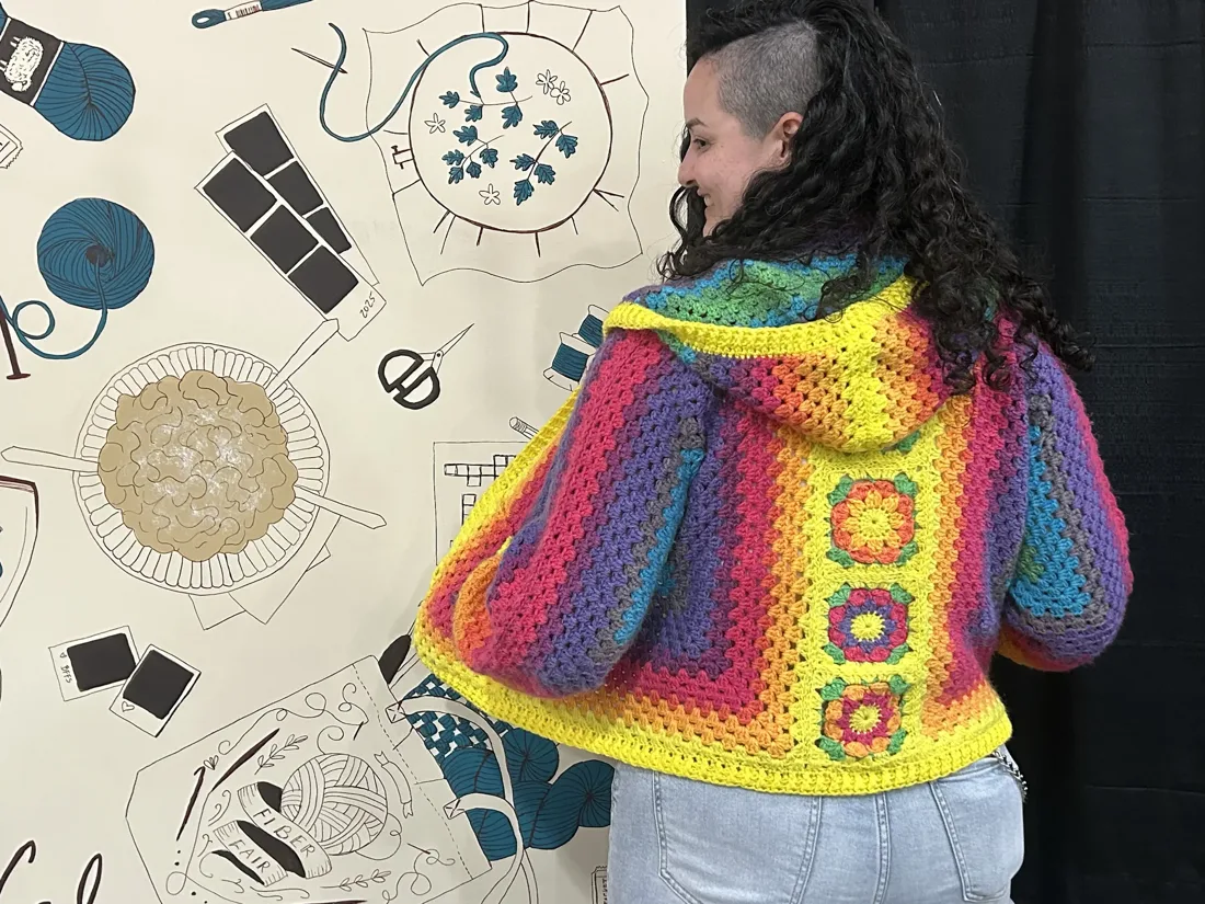 Anna Mendez Perdomo at the SoCal Fiber Fair, showing the back of a rainbow granny-square hoodie she crocheted